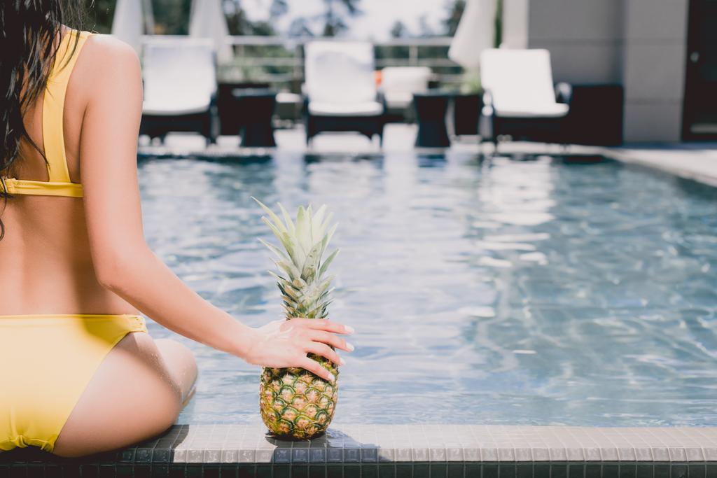 abgeschnittene Ansicht einer Frau im Badeanzug, die am Pool sitzt und reife Ananas in der Hand hält - Foto, Bild