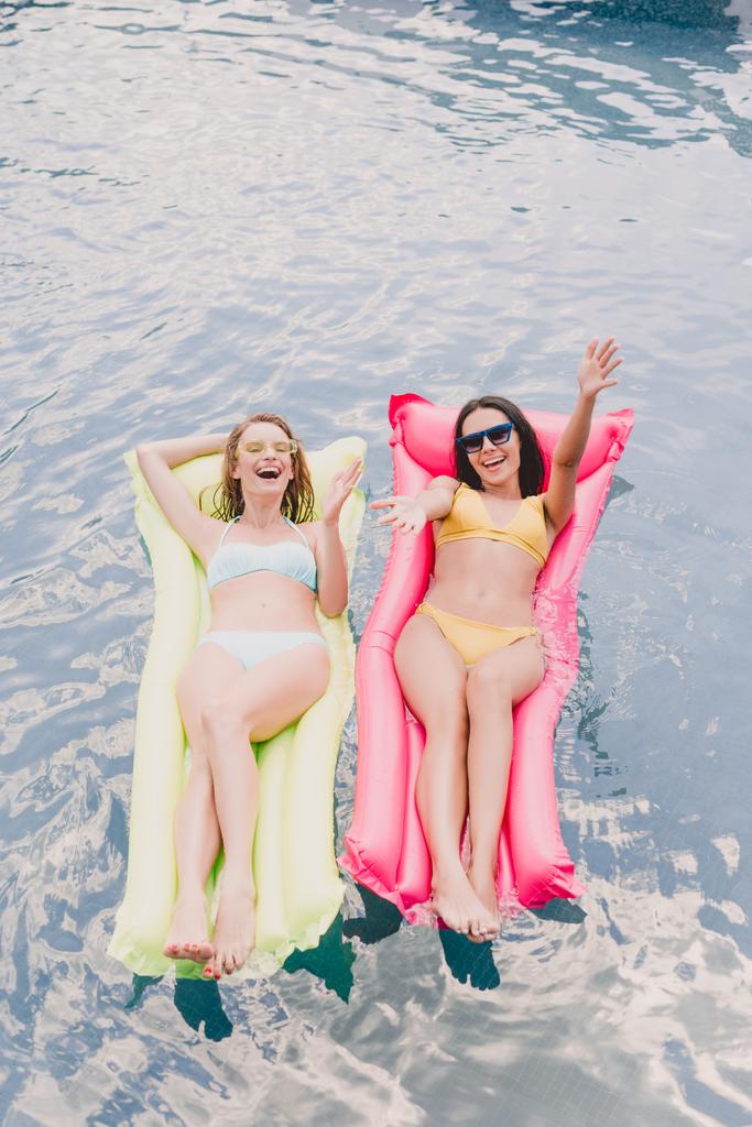 bovenaanzicht van brunette en blonde gelukkige en opgewonden vrouwen liggend op zwembad drijft met uitgestrekte handen in het zwembad - Foto, afbeelding