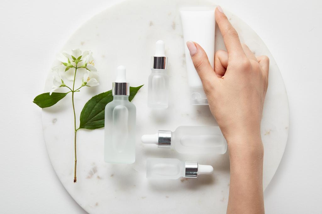 vista recortada de la mano de la mujer con tubo de crema sobre la placa con jazmín y botellas de vidrio cosmético en la superficie blanca
 - Foto, Imagen