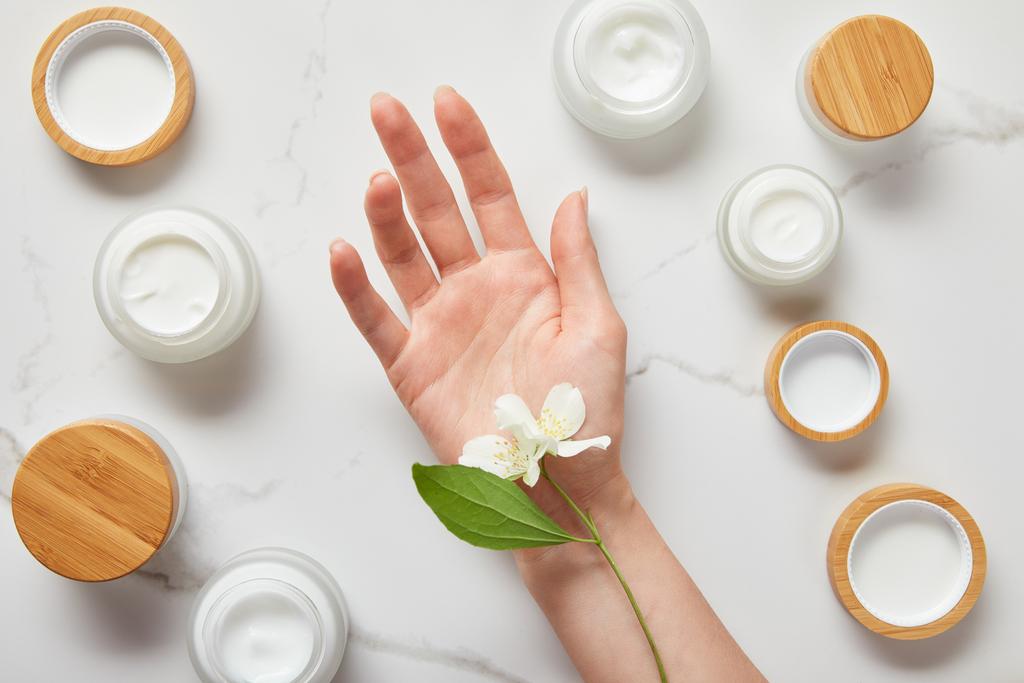 cropped view of jasmine flowers on woman hand near jars with cosmetic cream on white surface - Photo, Image