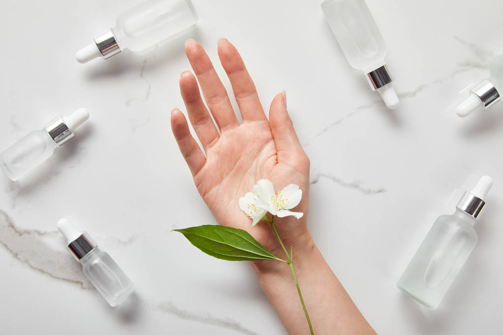 bijgesneden beeld van vrouw hand met jasmijn in de buurt van cosmetische glazen flessen op wit oppervlak - Foto, afbeelding