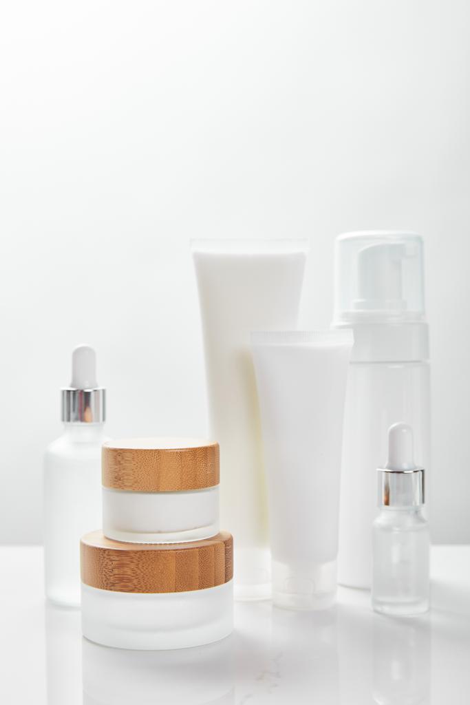 біла поверхня зі скляними пляшками, вершками в трубках, баночках та косметичному дозаторі
 - Фото, зображення