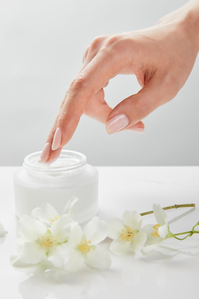 cropped view of woman hand touching cream in jar near jasmine flowers on white surface - Photo, Image