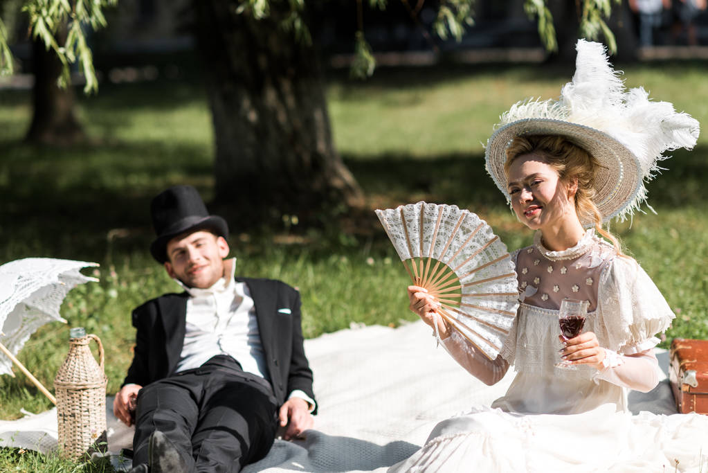 selektiver Fokus der glücklichen viktorianischen Frau, die auf einer Decke sitzt und ein Weinglas neben dem Mann hält  - Foto, Bild