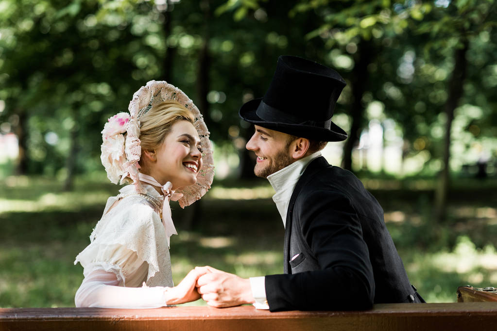 knappe aristocratische man die handen vasthoudt met vrolijke Victoriaanse vrouw in hoed  - Foto, afbeelding