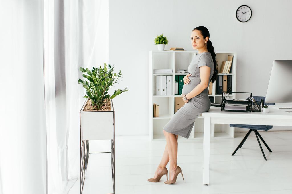 pohled na těhotnou ženu stojící v kanceláři s květináčem s rostlinným, stolním a knihovním případem - Fotografie, Obrázek