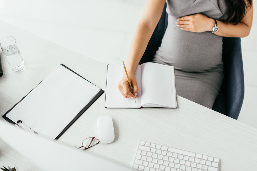 Ausgeschnittene Ansicht einer schwangeren Frau, die Notizen im Notizbuch macht, während sie im Büro hinter einem Tisch mit Tastatur, Ordner und Glas mit Wasser sitzt - Foto, Bild