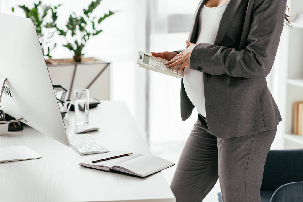 περικομμένη προβολή της εγκύου γυναίκα υπολογίζοντας ενώ στέκεται κοντά στο τραπέζι με τον υπολογιστή και το σημειωματάριο στο γραφείο - Φωτογραφία, εικόνα