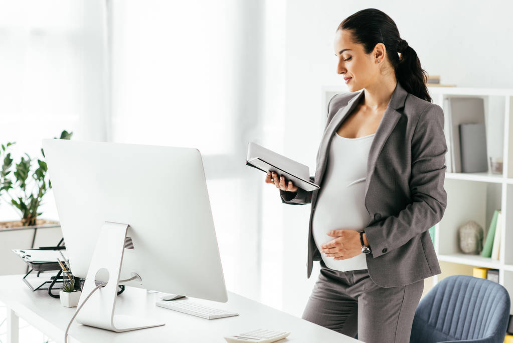 donna incinta che legge gli appunti dal taccuino mentre in piedi in ufficio vicino al tavolo con il computer
 - Foto, immagini