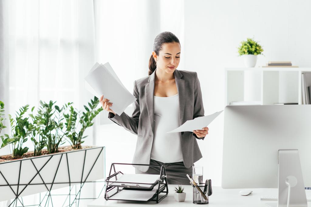  pregnant woman reading papers while standing in office near flowerpot and table with computer, document tray and pencil box - Photo, Image