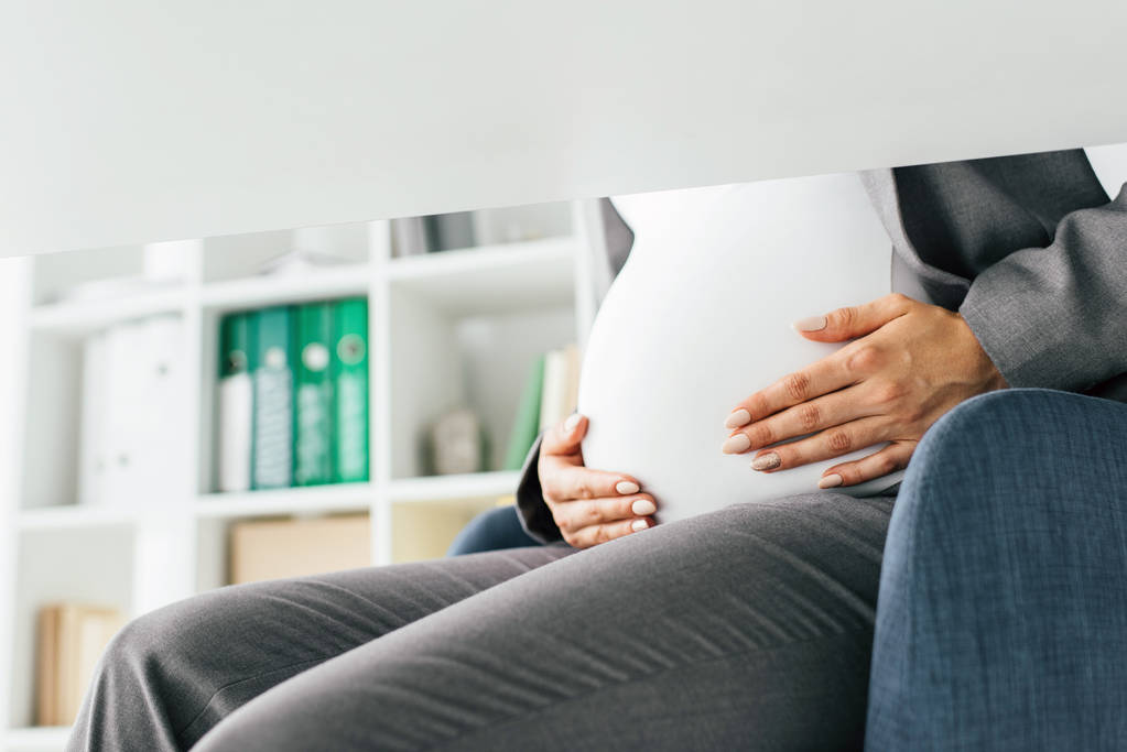 περικομμένη όψη της εγκύου γυναίκα που κρατά την κοιλιά ενώ κάθεται σε καρέκλα γραφείου  - Φωτογραφία, εικόνα