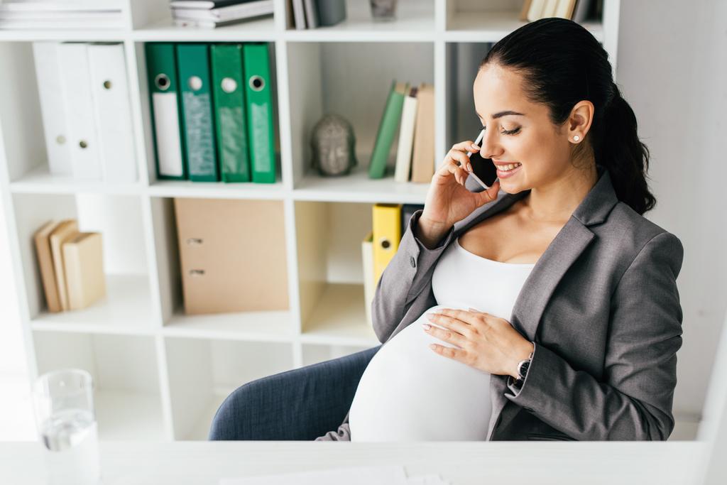 беременная женщина сидит за столом в офисе, разговаривает на смартфоне и ласкает живот
 - Фото, изображение