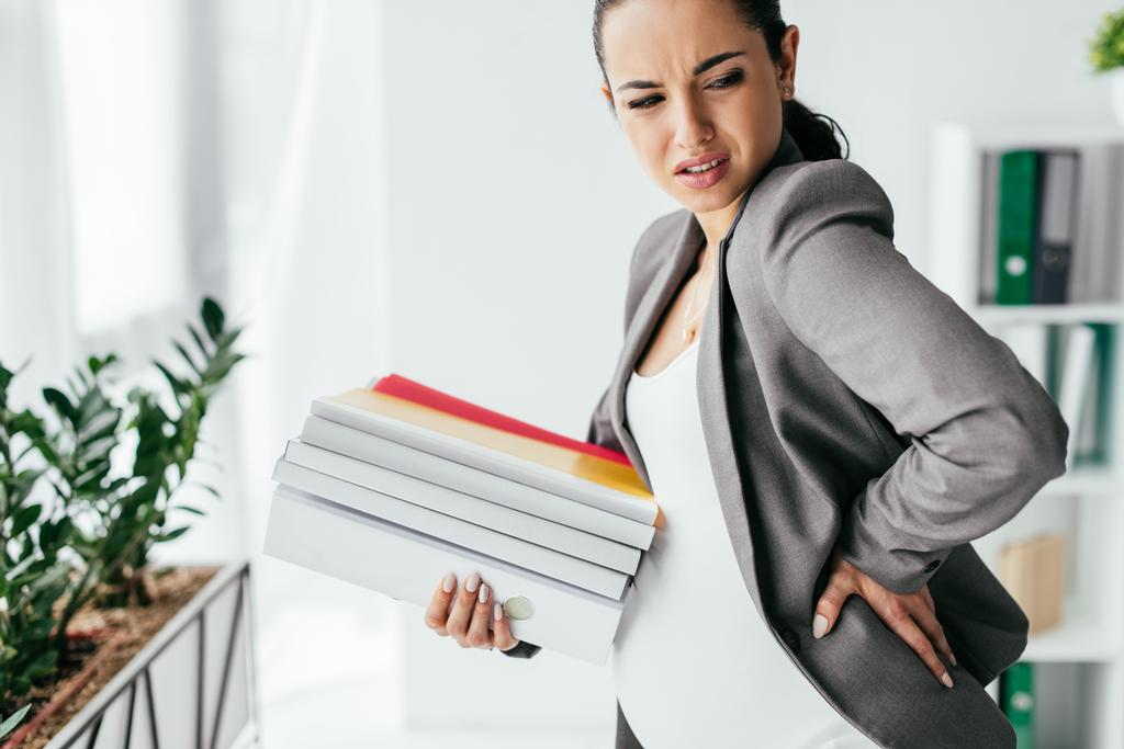 zwangere vrouw blijvende pijn en het houden van mappen terwijl staan in Office - Foto, afbeelding