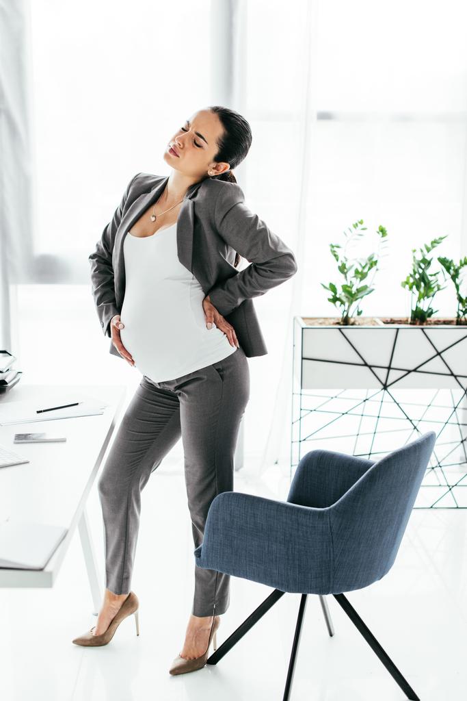 вигляд на повну довжину вагітної жінки, що стоїть біля офісного крісла в офісі і стійкий біль
 - Фото, зображення