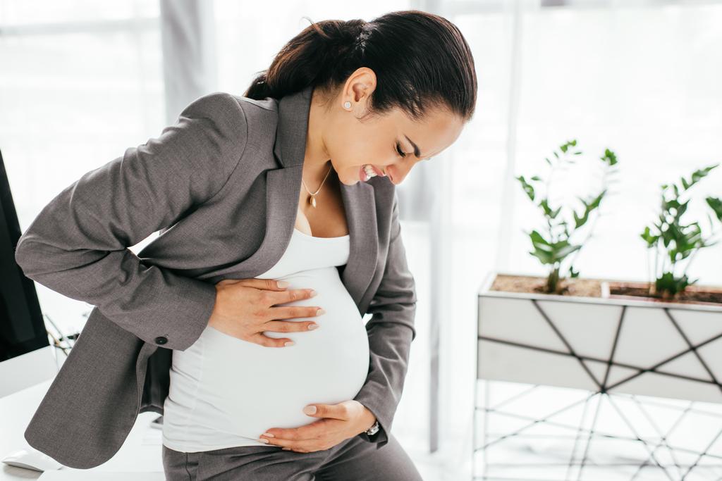 έγκυος γυναίκα που κρατά την κοιλιά και να κάνει γκρίσσος λόγω του πόνου, ενώ κάθονται στο γραφείο - Φωτογραφία, εικόνα