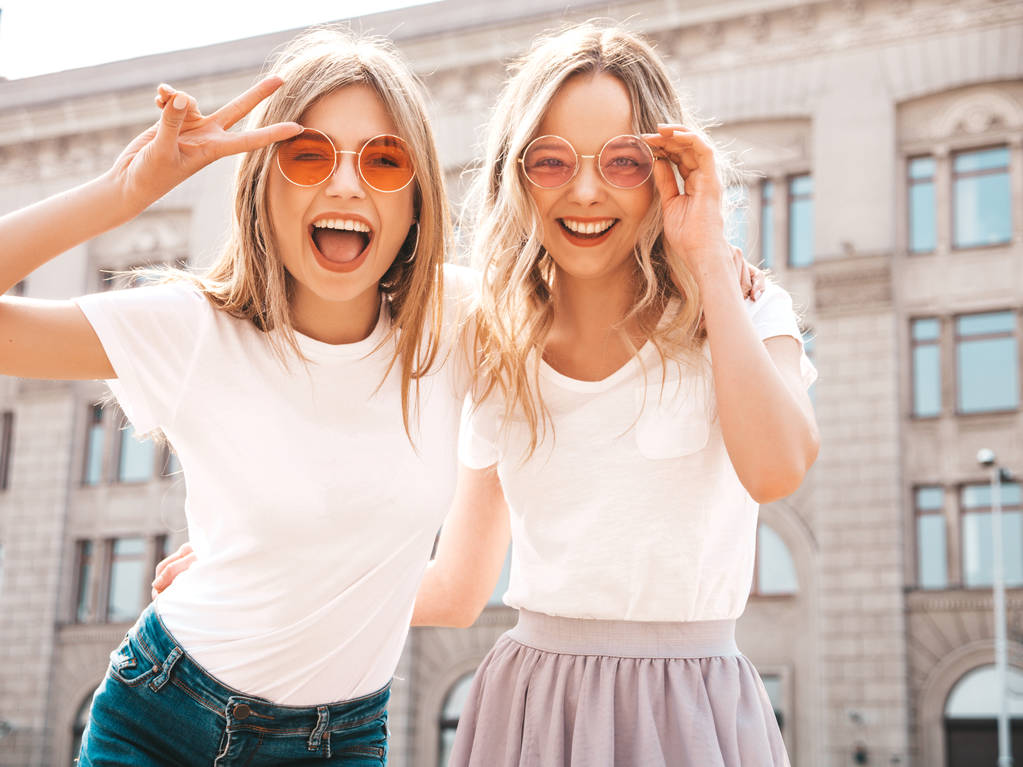 Porträt zweier junger hübscher blonder lächelnder Hipstermädchen in trendiger sommerweißer T-Shirt-Kleidung. sexy unbeschwerte Frauen posieren auf der Straße Hintergrund. positive Models, die Spaß an Sonnenbrillen haben. - Foto, Bild