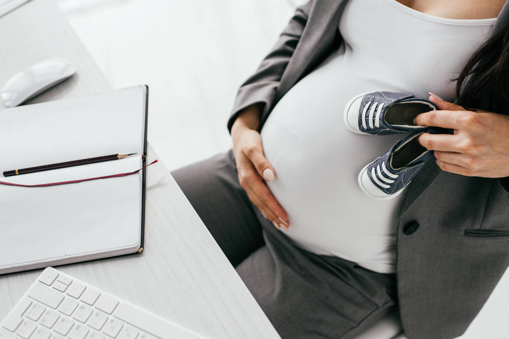 vista cortada de mulher grávida segurando pequenas botas de bebê enquanto sentado atrás da mesa com teclado e notebook
 - Foto, Imagem