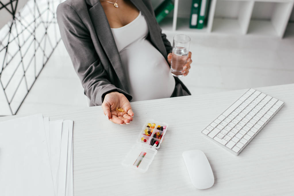 オフィスに座っている間、水で錠剤とガラスを持つ妊娠中の女性のトリミングされたビュー - 写真・画像