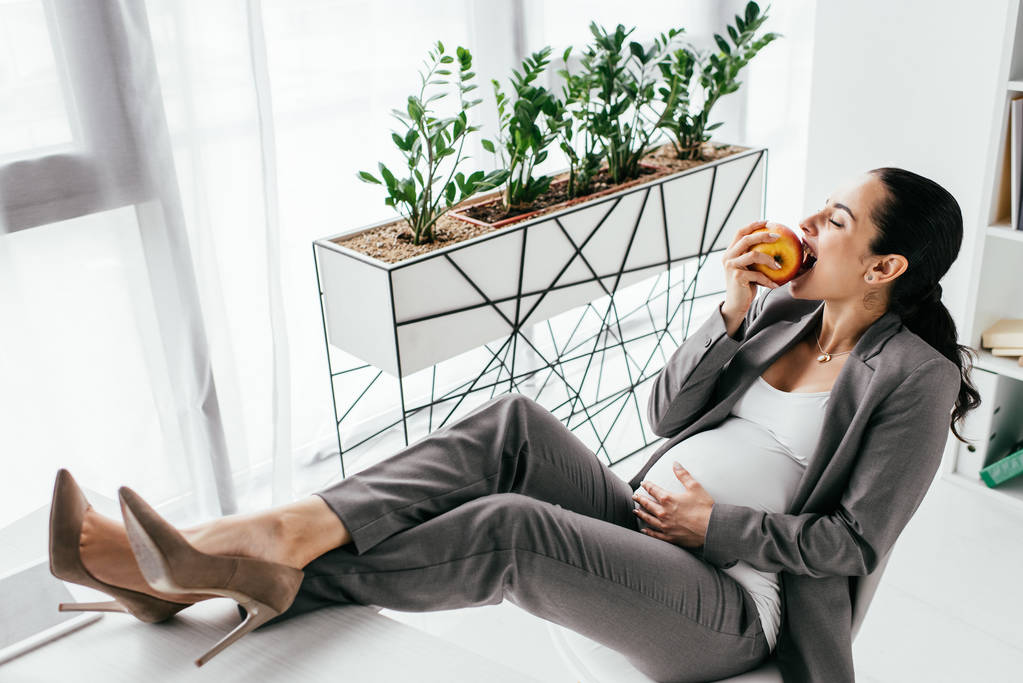 raskaana oleva nainen istuu työtuolissa ja syö omenaa
 - Valokuva, kuva