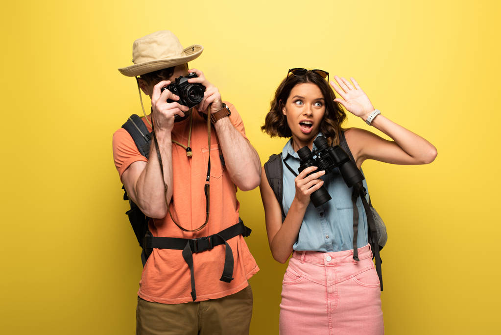 man het nemen van foto met digitale camera terwijl staande in de buurt van jonge vrouw zwaaien hand en kijken weg op gele achtergrond - Foto, afbeelding