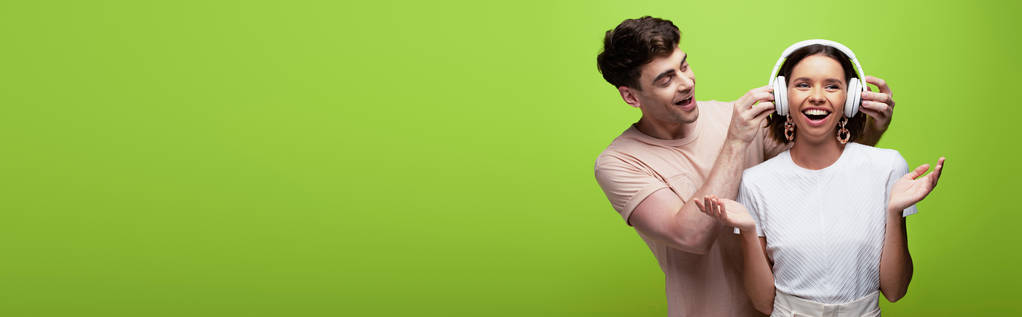 panoramic shot of handsome man putting on headphones on cheerful girl on green background - Photo, Image