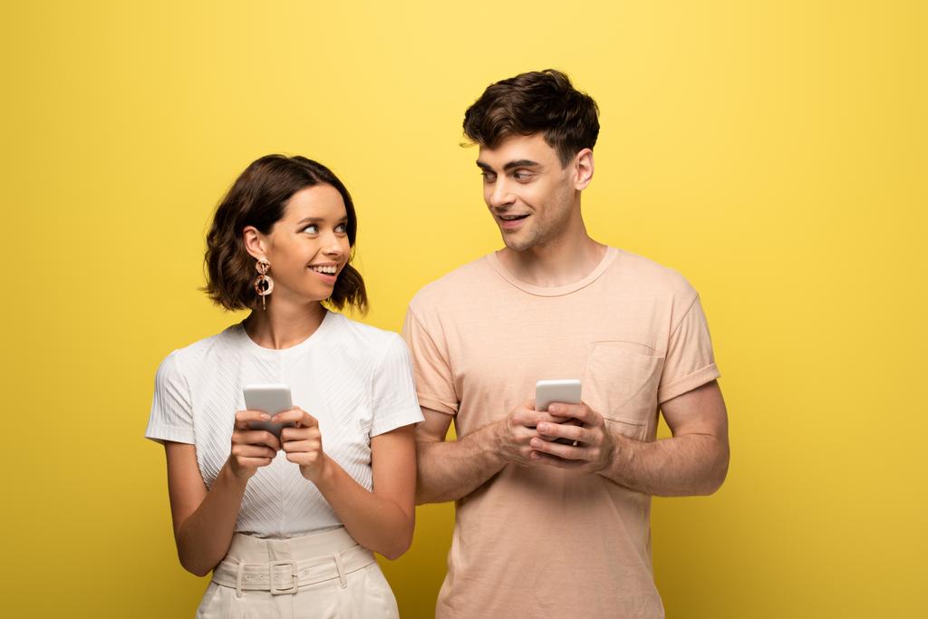 hombre y mujer positivos que se miran entre sí mientras usan teléfonos inteligentes sobre fondo amarillo
 - Foto, Imagen