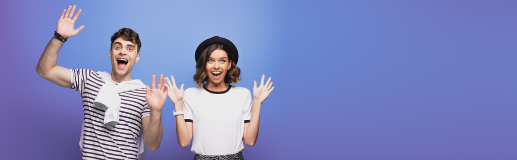 panoramic shot of excited man and woman waving hand and smiling at camera on blue background - Photo, Image