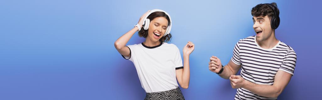 panoramische foto van opgewonden man en vrouw dansen tijdens het luisteren van muziek in een koptelefoon op een blauwe achtergrond - Foto, afbeelding