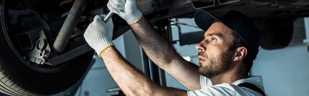 panoramic shot of  bearded car mechanic in cap repairing automobile in car service  - Photo, Image