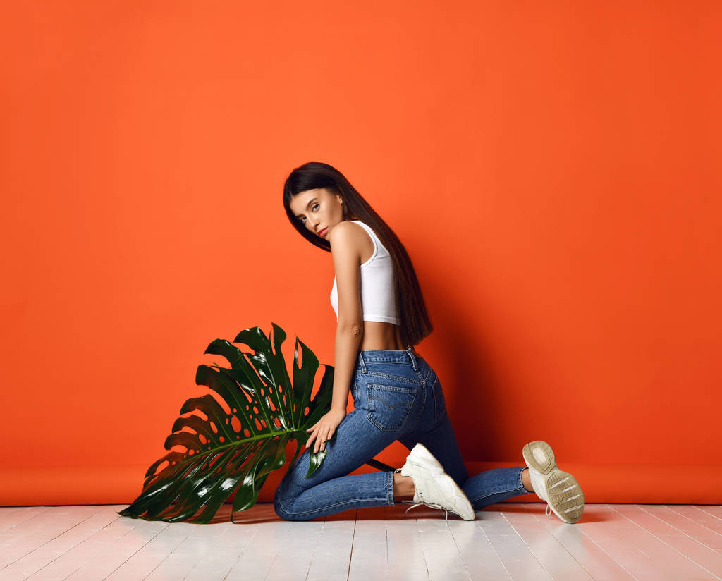 Молода красива дівчина хіпстера в джинсах і білому верхньому тримає великий тропічний лист на помаранчевому фоні
 - Фото, зображення