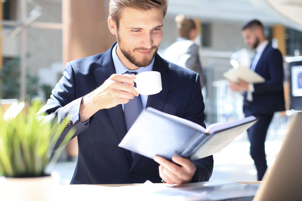 joven hombre de negocios beber café en la oficina mientras revisa portátil
. - Foto, Imagen