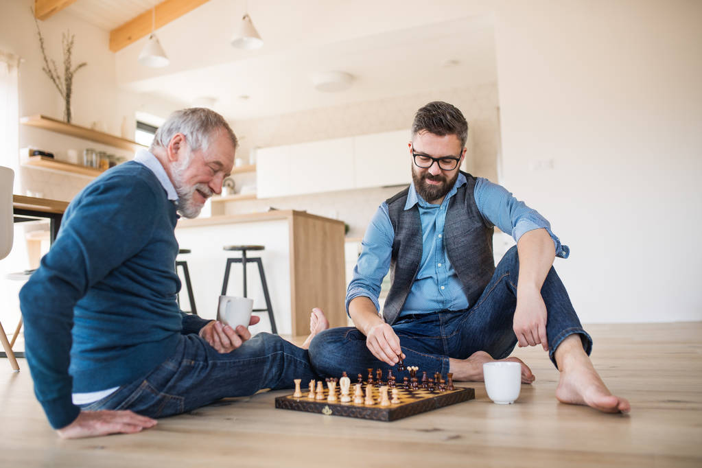An adult hipster son and senior father sitting on floor indoors at home, playing chess. - Photo, Image