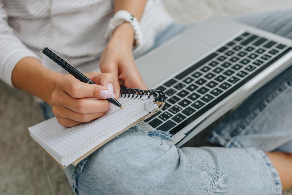 visão recortada da mulher escrevendo em notebook e segurando laptop
  - Foto, Imagem