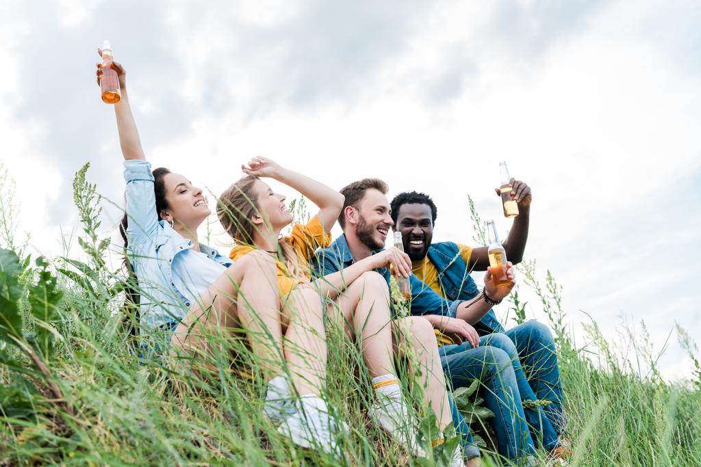 вибірковий фокус щасливих мультикультурних друзів, посміхаючись сидячи на траві і тримаючи пляшки з пивом
  - Фото, зображення