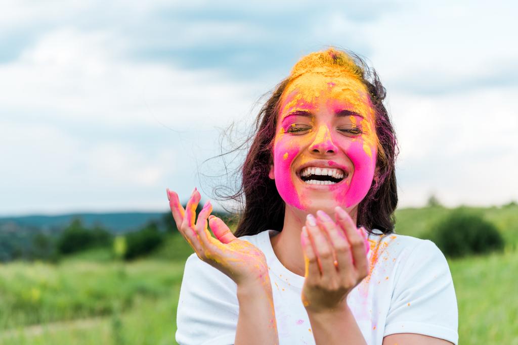 happy woman standing with closed eyes and pink and yellow holi paint on face  - Photo, Image