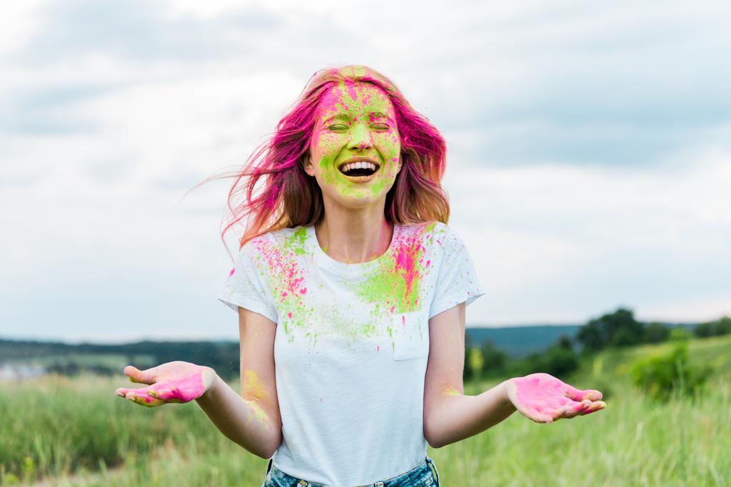 mulher alegre com olhos fechados e pintura holi rosa no rosto gesticulando e sorrindo ao ar livre
  - Foto, Imagem