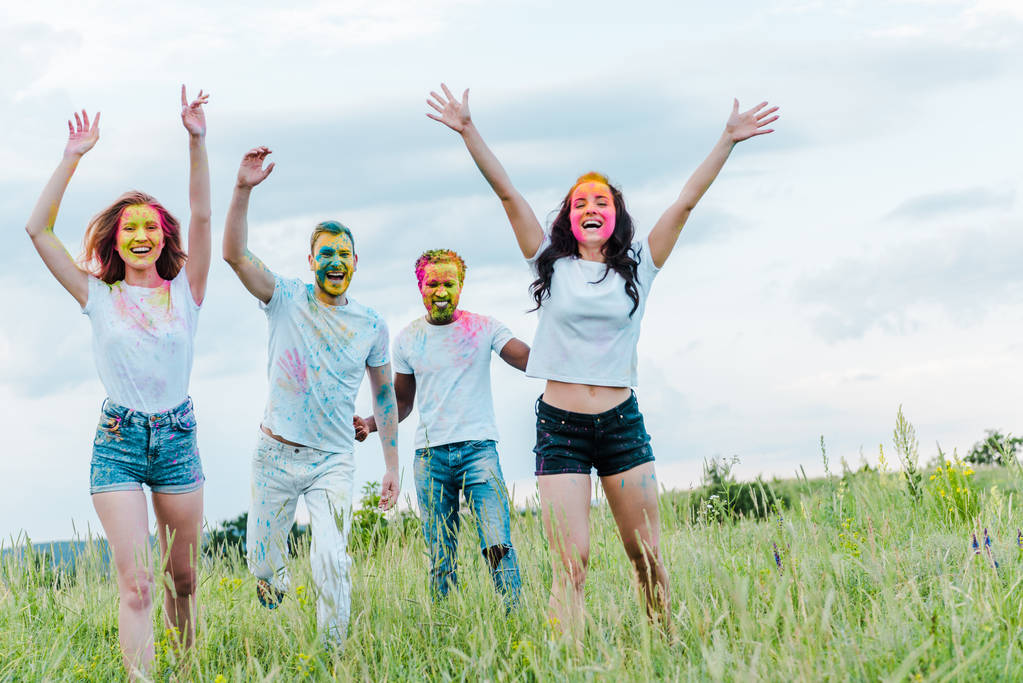 fröhliche multikulturelle Freunde mit bunten Holi-Farben auf Gesichter gestikulierend, während sie draußen stehen  - Foto, Bild
