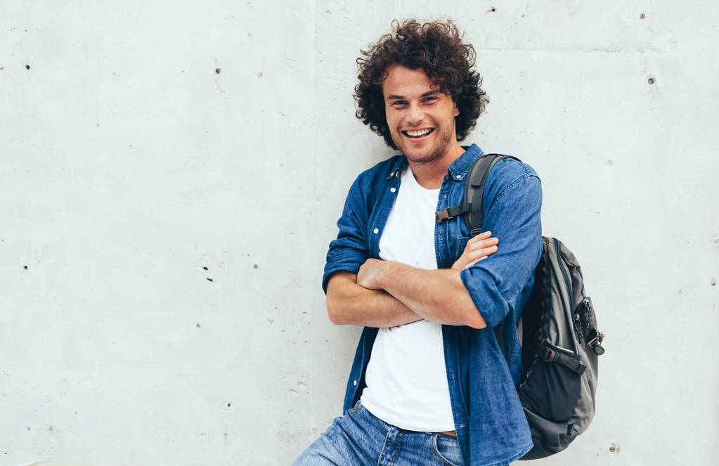 Pohledný úsměv mladého muže s batohu na zádech, stojící na stavbě betonového zázemí na městské ulici. Nezávislý muž s příležitostným oblečem působícím venku. Lidé a životní styl. - Fotografie, Obrázek