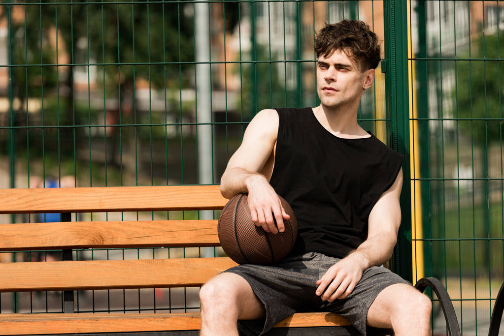 serious basketball player with ball sitting on wooden bench in sunny day at basketball court  - Photo, Image