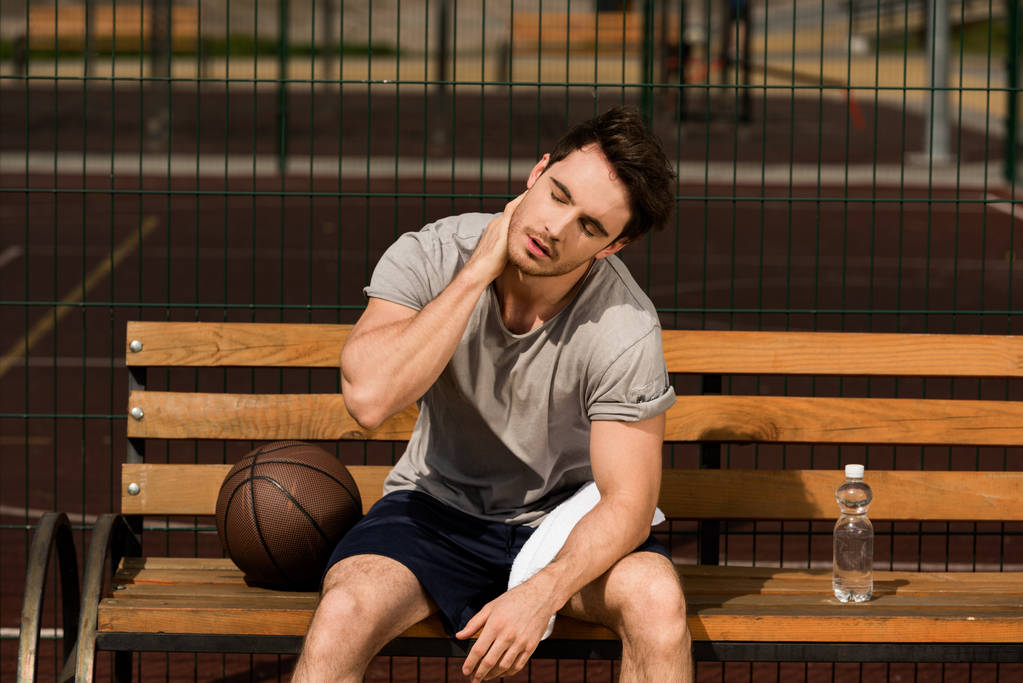 basketbalspeler zittend op houten bank en handdoek en fles water bij basketbalveld  - Foto, afbeelding