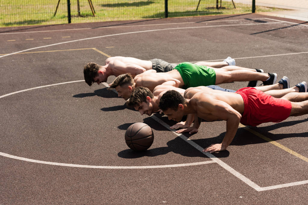 vier sexy Sportler ohne Hemd machen Liegestütze auf dem Basketballplatz bei sonnigem Tag - Foto, Bild