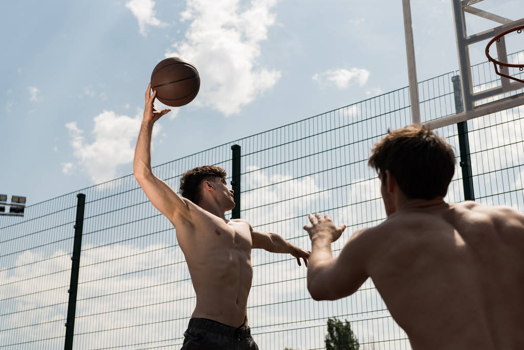 バスケットボールコートで青空の下でバスケットボールをしている2人のセクシーなシャツレススポーツマン - 写真・画像