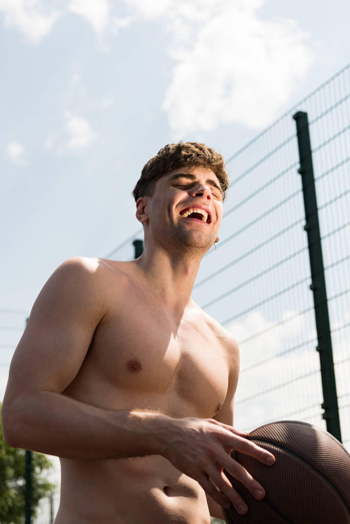 улыбающийся сексуальный спортсмен без рубашки держит мяч на баскетбольной площадке под голубым небом
 - Фото, изображение