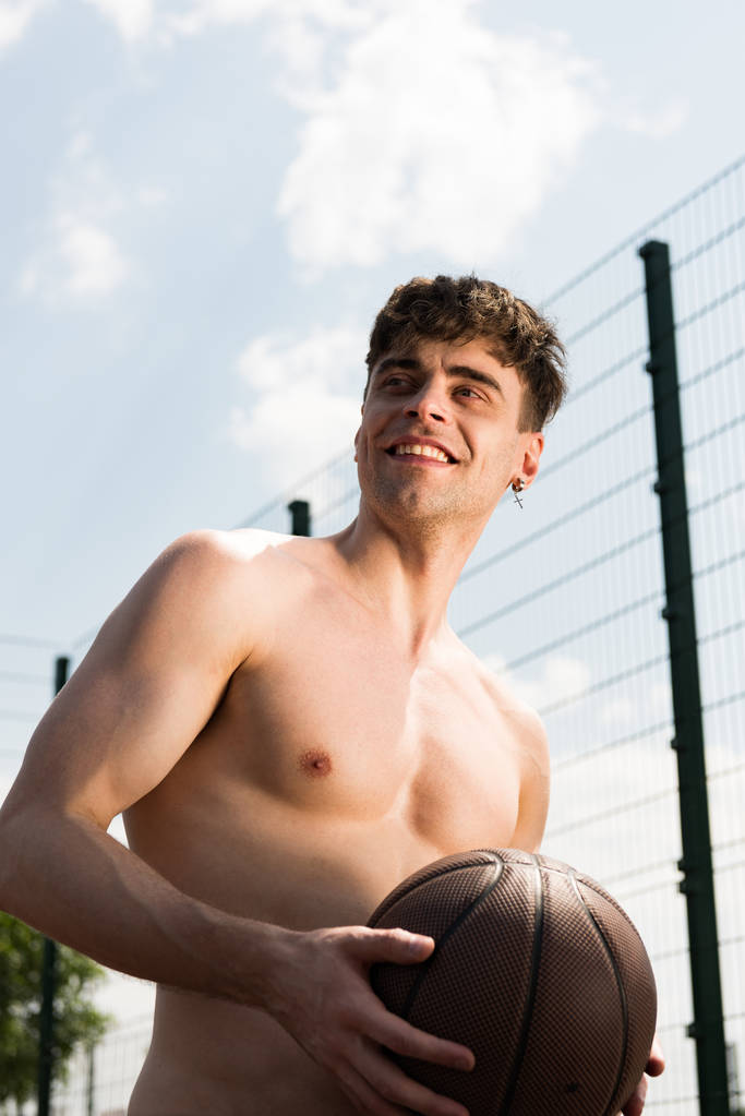 усміхнений сексуальний спортсмен без сорочки тримає м'яч на баскетбольному майданчику під блакитним небом
 - Фото, зображення