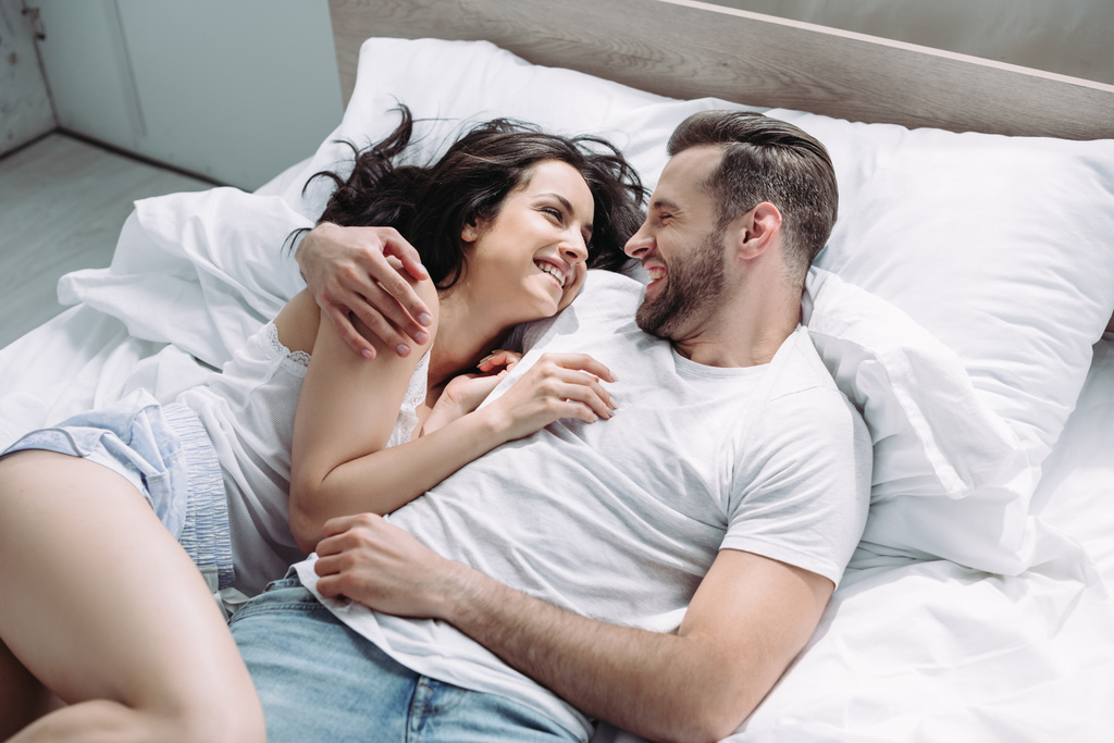 ελκυστική και μελαχρινή γυναίκα και ο άνθρωπος χαμογελά, ξαπλωμένοι στο κρεβάτι και αγκαλιάζοντας  - Φωτογραφία, εικόνα