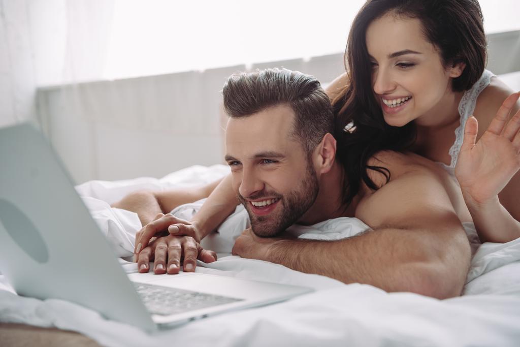 beautiful woman waving and handsome man using laptop in bedroom  - Photo, Image