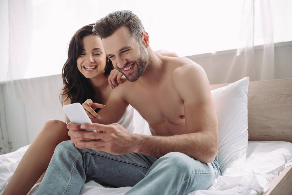 hombre guapo y sonriente sosteniendo el teléfono inteligente y la mujer señalándolo con el dedo
  - Foto, Imagen