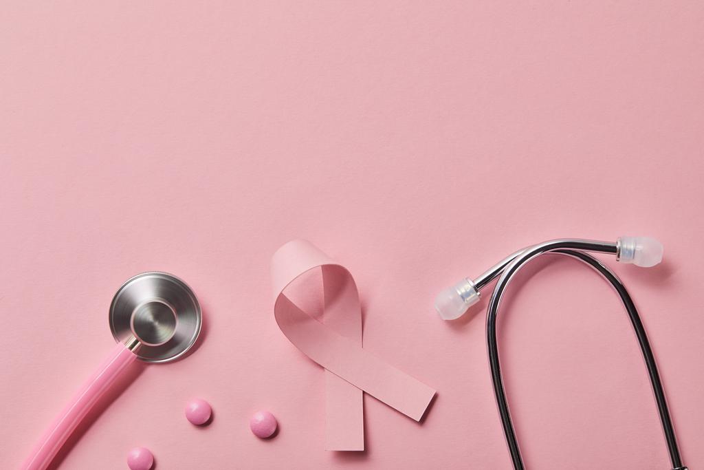 Draufsicht auf rosa Metall-Stethoskop, Brustkrebsband und drei Pillen auf hellrosa Hintergrund - Foto, Bild