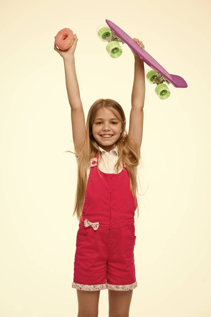 Kid girl relax hold penny board. Active leisure concept. Girl raise up penny board and sweet donut on white background. Originally designed girls skateboard. Plastic bright penny board for children - Photo, Image
