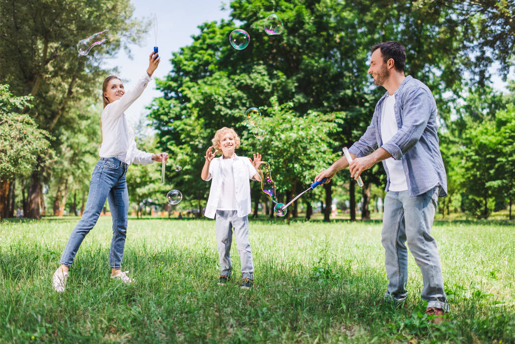 mother, father and son in casual clothes playing with soap bubbles in park - Photo, Image